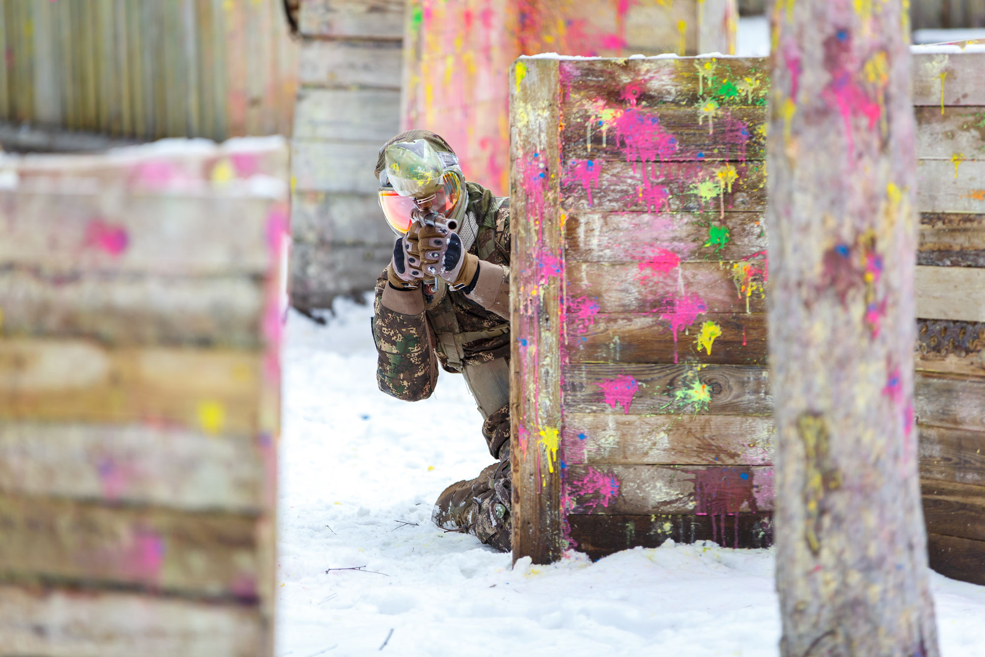 Gracz paintballa w kamuflażu celuje zza drewnianej osłony na zaśnieżonym polu