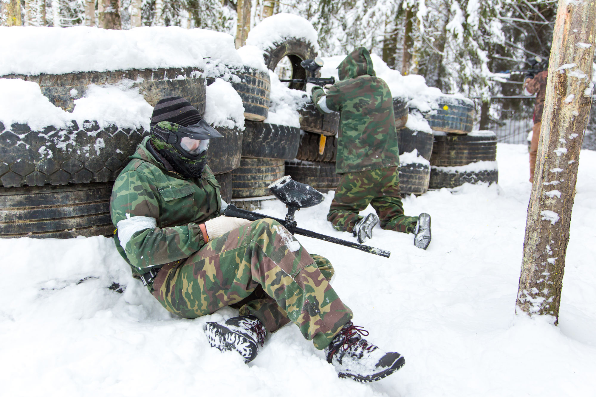 Dwóch graczy paintballa w zimowym lesie, w kamuflażu, za osłoną z opon pokrytych śniegiem