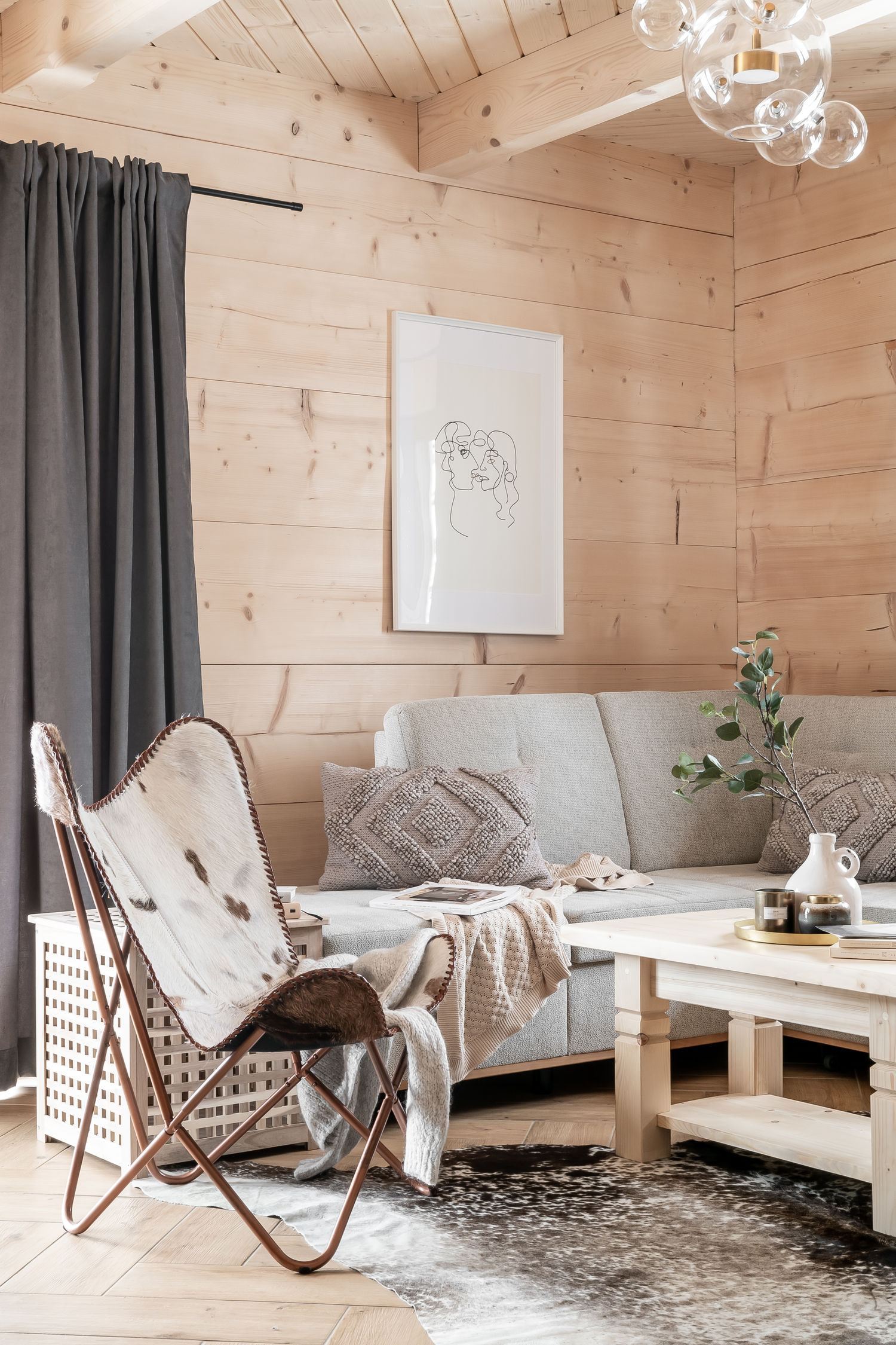 Przytulny salon z drewnianymi ścianami, jasną sofą, fotelami, stolikiem i czarnymi zasłonami